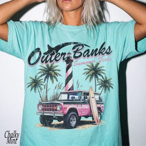 May include: A light blue t-shirt with a graphic featuring the text "Outer Banks Summer Feels". The design includes a pink and black SUV, a striped lighthouse, palm trees, and a surfboard. The shirt has a relaxed fit.