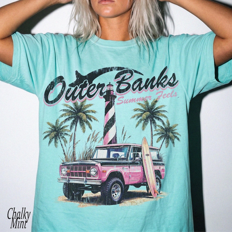 May include: A light blue t-shirt with a graphic featuring the text "Outer Banks Summer Feels". The design includes a pink and black SUV, a striped lighthouse, palm trees, and a surfboard. The shirt has a relaxed fit.