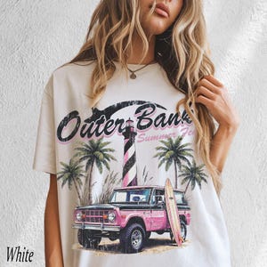 May include: White t-shirt with a graphic design featuring the text "Outer Banks Summer Fest". The design includes a pink and black striped lighthouse, palm trees, a pink SUV, and a surfboard. The word "White" is printed at the bottom left.