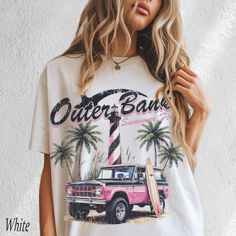 May include: White t-shirt with a graphic design featuring the text "Outer Banks Summer Fest". The design includes a pink and black striped lighthouse, palm trees, a pink SUV, and a surfboard. The word "White" is printed at the bottom left.