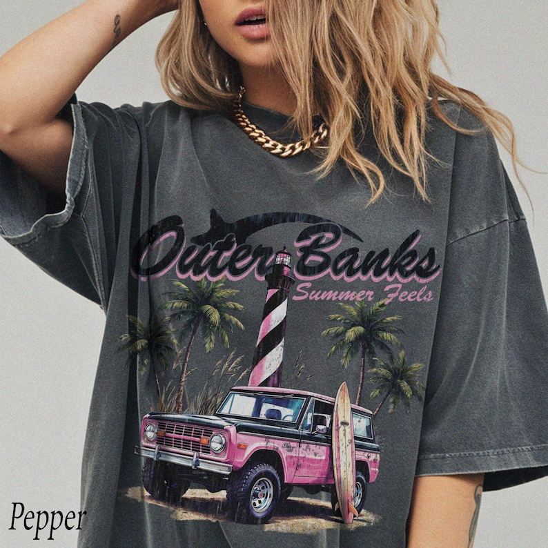 May include: Dark gray t-shirt with a graphic featuring the text "Outer Banks Summer Feels." The design includes a pink SUV, a surfboard, a lighthouse, and palm trees. The word "Pepper" is printed at the bottom left.