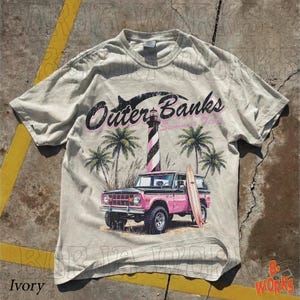 May include: Ivory-colored t-shirt featuring a graphic of the Outer Banks with a pink and black SUV, a surfboard, palm trees, and a lighthouse. The text "Outer Banks Summer 2023" is printed above the image.