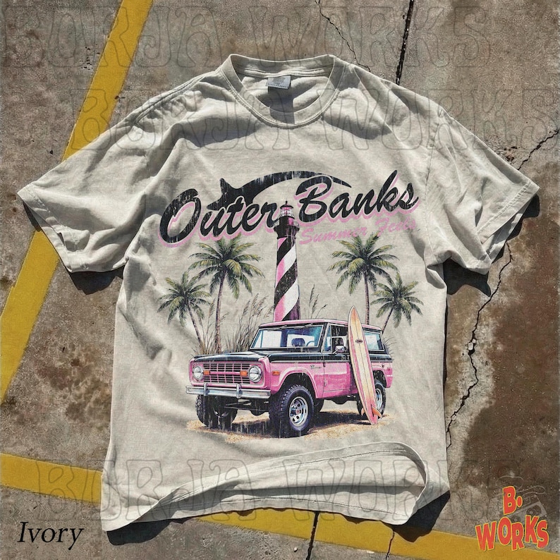 May include: Ivory-colored t-shirt featuring a graphic of the Outer Banks with a pink and black SUV, a surfboard, palm trees, and a lighthouse. The text "Outer Banks Summer 2023" is printed above the image.