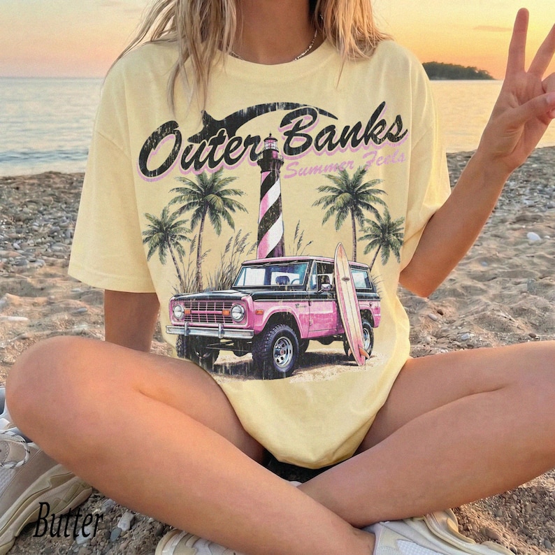 May include: Butter-colored t-shirt with a graphic featuring the text "Outer Banks Summer Feels", a pink SUV, a surfboard, palm trees, and a lighthouse. The shirt is worn by a person sitting on a sandy beach.
