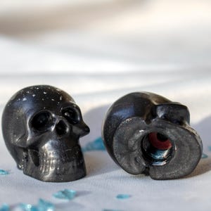 May include: Two black skull-shaped objects, one upright and one on its side, against a white backdrop. The upright skull has a speckled surface. The other skull reveals a red interior. Blue glass shards are scattered in the foreground.