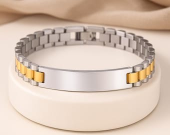 May include: A stainless steel bracelet with gold-coloured accents. The bracelet features a silver-coloured centre plate and a chain-link design with alternating silver and gold-coloured links. The bracelet is displayed on a white surface.