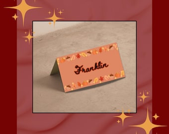 May include: A folded, peach-colored place card with the name "Franklin" in black script. The card is decorated with a border of fall leaves and pumpkins. The background is a neutral gray surface, with a dark red border and gold starburst accents.
