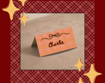 May include: A folded place card with the name "Charlie" in cursive script. The card has a peach and white checkered pattern and a black bow illustration. The background includes a red plaid border and gold star accents.