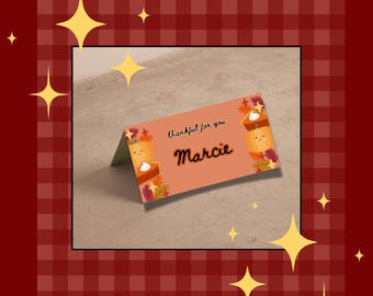 May include: A folded, peach-colored place card with the text "thankful for you" and the name "Marcie" in cursive. The card features illustrations of pumpkins, pie, and fall leaves. The background is a red and brown plaid pattern.