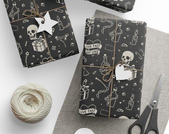 May include: Several wrapped gifts with black paper featuring white skull and gift box illustrations, tied with twine and adorned with tags. The phrase "LIVE FAST EAT CAKE" is printed on the wrapping. A ball of twine, scissors, and blank tags are also present.