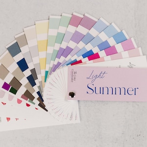 Pode incluir: Um leque de paleta de cores com várias tonalidades, incluindo rosas, azuis, verdes e neutros. O leque está rotulado como "Light Summer" e é projetado para análise de cores. A paleta está disposta em semicírculo, mostrando uma gama de tons.