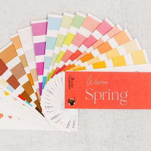 Może przedstawiać: Wachlarz palety kolorów z różnymi kolorami, w tym różowym, pomarańczowym, żółtym i niebieskim. Dołączona jest czerwona karta z napisem "Warm Spring". Widoczne jest również logo The Color Coordinator.