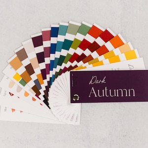 Puede incluir: Un abanico de paleta de colores con una variedad de tonos, incluyendo tonos marrones, naranjas, verdes y azules. El abanico está etiquetado como "Dark Autumn" en escritura blanca sobre una tarjeta morada oscura. Sobre una superficie blanca.
