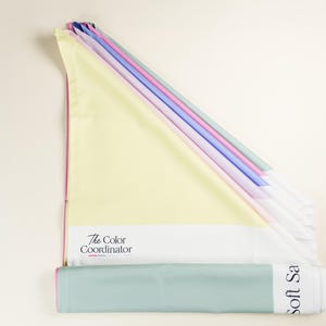 May include: A collection of pastel-colored fabric swatches, including yellow, pink, blue, and green. The swatches are arranged in a fan shape, with a rolled-up section at the bottom. Text on the fabric reads "The Color Coordinator" and "Soft Sa".