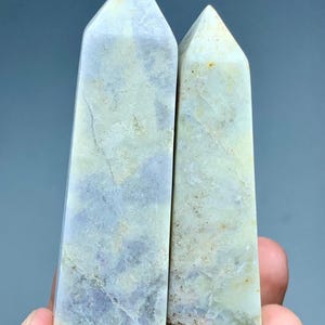 Peut inclure: Deux obélisques en cristal poli. Les obélisques sont rectangulaires avec un sommet pointu. Les cristaux sont de couleur lavande pâle et crème avec des veines naturelles. Les obélisques sont tenus dans une main.