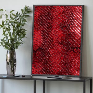 May include: A framed art print with a close-up of a red snakeskin pattern. The artwork has a glossy finish and is set in a black frame. The print is displayed on a dark grey table with a vase of green leaves.