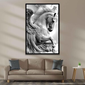 May include: Black and white framed print of a sculpted Pegasus, a mythical winged horse. The artwork features detailed wings and flowing mane, set against a bright background. The print is displayed above a beige sofa with blue pillows.
