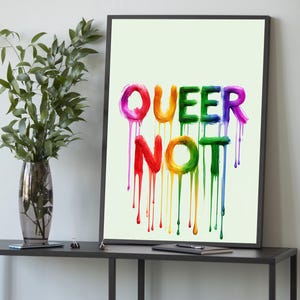 May include: A framed art print with the words "QUEER NOT" in rainbow colors. The letters appear to be painted with dripping paint, creating a vibrant and eye-catching design. The artwork is displayed on a black table.