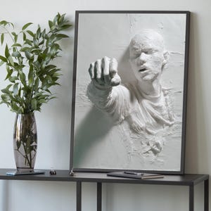 Könnte beinhalten: Gerahmte weiße Reliefskulptur einer Person, die mit geballter Faust nach vorne zeigt. Das Kunstwerk ist vor einem weißen Hintergrund und in einem dunkelgrauen Rahmen eingefasst. Die Skulptur steht auf einem dunkelgrauen Tisch mit einer Vase mit grünen Blättern.