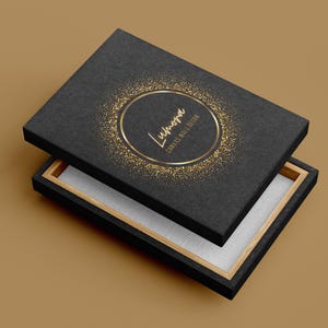 May include: Black box with a gold glitter design and the word "Lumora" in gold lettering. The box is open, revealing a white interior and a wooden frame. The words "Canvas Wall Design" are also visible.