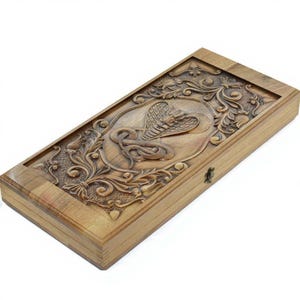 Könnte beinhalten: Rechteckige Holzbox mit einem Relief einer Kobra. Der Deckel ist mit aufwendigen floralen Mustern und einem zentralen ovalen Rahmen verziert. Die Box ist aus hellbraunem Holz gefertigt und hat einen kleinen Metallverschluss an der Seite.