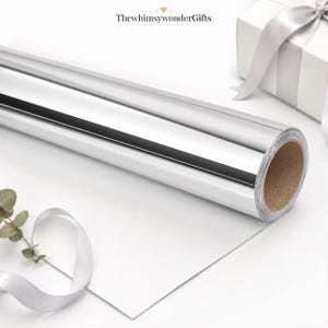 May include: A roll of reflective silver wrapping paper with a smooth metallic finish and a brown cardboard core. The text ThewhimsywonderGifts is printed at the top. A white gift box with a silver ribbon and a sprig of green eucalyptus are visible.