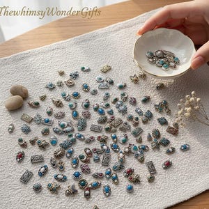 May include: An assortment of small, decorative metal embellishments with turquoise, red, and white details, displayed on a white surface. Some are in a small, white, scalloped dish. The text "ThewhimsyWonderGifts" is visible.