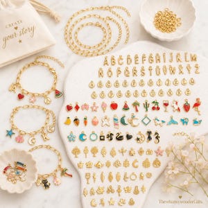 May include: A collection of gold-colored jewelry charms and bracelets. The charms include letters, fruits, animals, and other shapes. A small bag with the text "CREATE your story" is also visible.