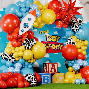 May include: A vibrant balloon display featuring a rocket, a red star, and the text "It's a Boy Story." The balloons are red, yellow, blue, and white with cow print patterns. Wooden blocks spell "ABC."