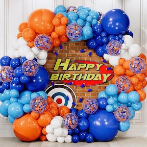 May include: A festive balloon arch in shades of blue, orange, and white, with confetti-filled balloons. The backdrop features a brick wall with a "Happy Birthday" sign and a target. A celebratory decoration.