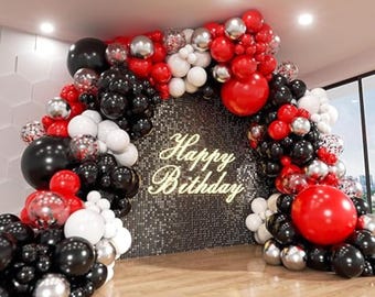 May include: A festive birthday archway made of black, red, silver, and white balloons. The arch frames a backdrop with the words "Happy Birthday" in gold script. The balloons are arranged in clusters, creating a celebratory atmosphere.