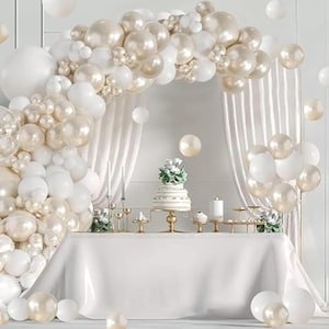 May include: A festive scene with a white tablecloth, a tiered cake, and an arch of white and gold balloons. The balloons create a backdrop for the table, with additional balloons scattered around the room. Candles and decorative elements complete the elegant party setting.