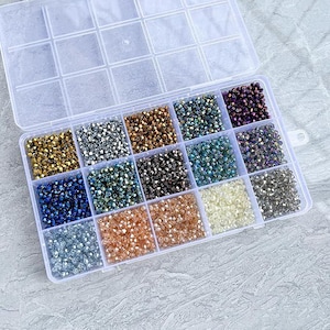 May include: A clear plastic organizer filled with various colors of small, faceted beads. The beads are arranged in separate compartments, showcasing a range of colors including gold, silver, blue, green, and purple. The organizer is open, displaying the contents.