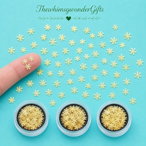 May include: A collection of small gold-colored metal snowflake-shaped charms scattered on a bright turquoise background, with three clear circular containers filled with the same snowflake charms and the text ThewhimsywonderGifts at the top.
