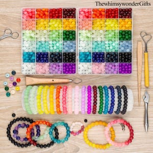 May include: A craft kit with colorful beads and bracelets. Two clear plastic organizers hold beads in various colors, including red, orange, yellow, green, blue, and purple. Several beaded bracelets in different colors are displayed, along with tools like scissors and a heart-shaped tool.