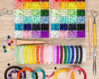 May include: A craft kit with colorful beads and bracelets. Two clear plastic organizers hold beads in various colors, including red, orange, yellow, green, blue, and purple. Several beaded bracelets in different colors are displayed, along with tools like scissors and a heart-shaped tool.