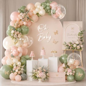 May include: A baby shower backdrop with a pastel color scheme. The display includes a balloon arch in pink, white, and green, with floral accents. A sign reads "Oh Baby" and another says "a little Butterfly is on the way."