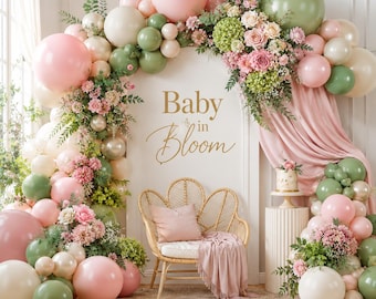 May include: A baby shower backdrop featuring a floral arch with pink, green, and white balloons. The arch frames a white wall with the words "Baby in Bloom" in gold script. A wicker chair with a pink pillow and blanket is in front.