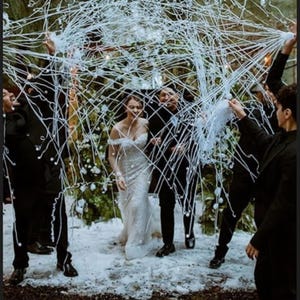 May include: A bride in a white, off-the-shoulder wedding dress walks through a shower of white streamers, surrounded by people in black suits. The scene takes place in a snowy, outdoor setting, with trees in the background.