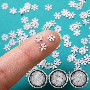 May include: Tiny, white snowflake-shaped embellishments scattered on a teal surface. Three snowflakes rest on a fingertip, with three small, round containers at the bottom. These decorative elements are ideal for crafts and winter-themed projects.