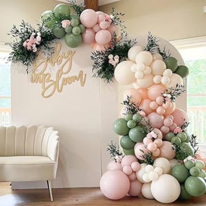 May include: A decorative archway with a balloon garland in shades of pink, green, and cream, accented with greenery and small pink flowers. The words "Baby in Bloom" are displayed in gold script. A white backdrop and a cream-coloured sofa complete the scene.