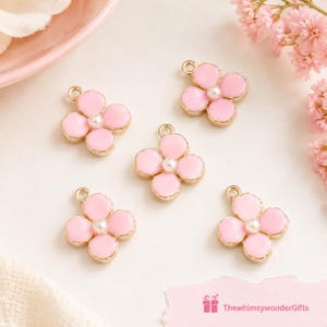 May include: Five flower-shaped charms with gold-toned metal edges, light pink enamel petals, and a small white faux pearl at the centre of each. Each charm features a small circular loop at the top for attachment. Text reads ThewhimsywonderGifts.