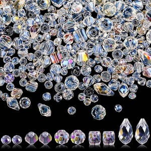 May include: A collection of clear, iridescent crystal beads in various shapes and sizes. The beads include round, faceted, and teardrop designs, reflecting light with a rainbow effect. The beads are scattered on a black background.