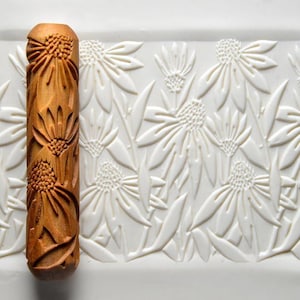 May include: A wooden rolling pin with a floral design. The pin is a warm brown color and features a repeating pattern of detailed flowers. The background is a white surface with a matching embossed floral pattern.