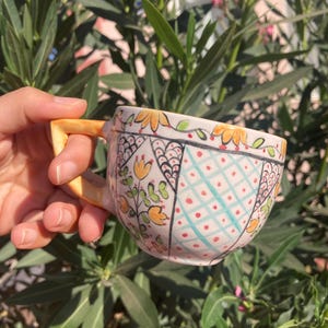 Può includere: Una tazza da tè in ceramica dipinta a mano con manico e bordo gialli. La tazza presenta un colorato design floreale e geometrico in arancione, blu e rosso. Lo sfondo è un cespuglio verde lussureggiante.
