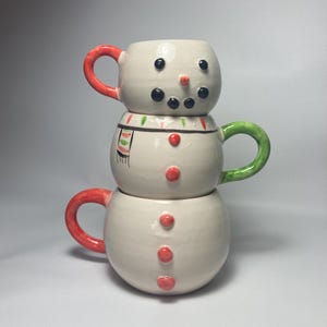 May include: Three-part snowman mugs with red, green, and black detailing. The top mug has a red handle, black eyes, and a carrot nose. The middle mug has a green handle and a scarf design. The bottom mug has a red handle and red buttons.