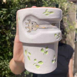 Peut inclure: Pot de fleurs en céramique blanche avec une illustration de girafe et des découpes de feuilles vertes. La girafe est peinte avec des détails beiges et bruns. Le bord est peint en orange.