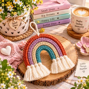 May include: A crocheted rainbow wall hanging featuring concentric arches in rust orange, lavender, beige, and teal, finished with cream-coloured fringe tassels at both ends and a braided hanging loop at the top, centred on a round wooden slice.