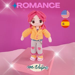 Pode incluir: Boneca de crochê com cabelo rosa, usando uma jaqueta laranja, calças cinzas com corações rosa e sapatos amarelos. A palavra "ROMANCE" está acima da boneca. A boneca está em uma superfície rosa. A marca "Mr. Vulpis" está na parte inferior.