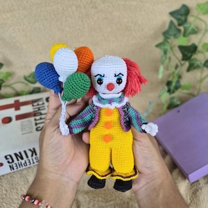 Pode incluir: Boneca de palhaço de crochê com cabeça branca, cabelo ruivo e uma roupa colorida. A boneca está segurando um monte de balões coloridos. O palhaço está usando um macacão amarelo com botões laranja e uma jaqueta listrada roxa, verde e preta.